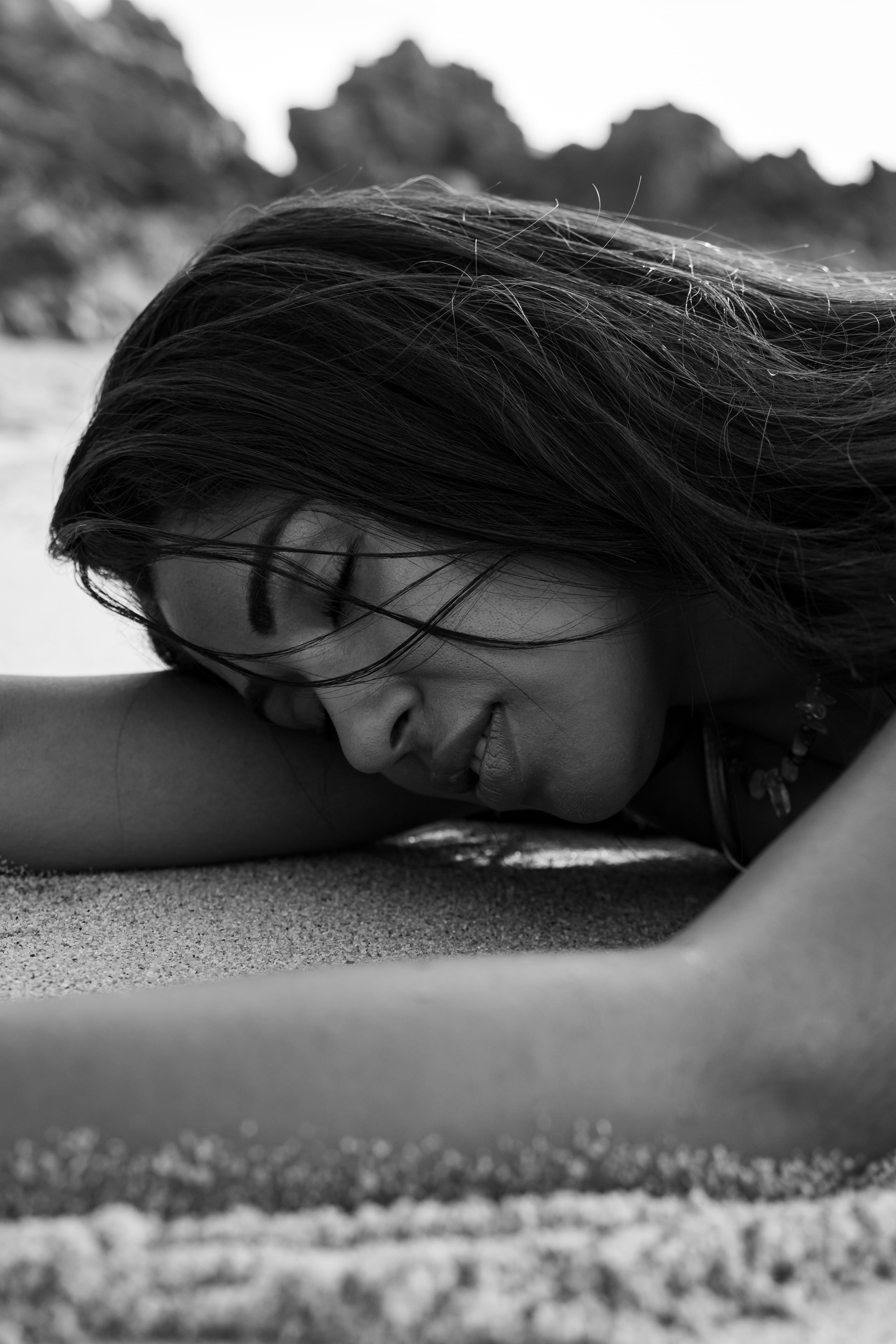 Schwarz-Weiß-Nahaufnahme einer Frau, die mit geschlossenen Augen und wehendem Haar im Sand ruht.