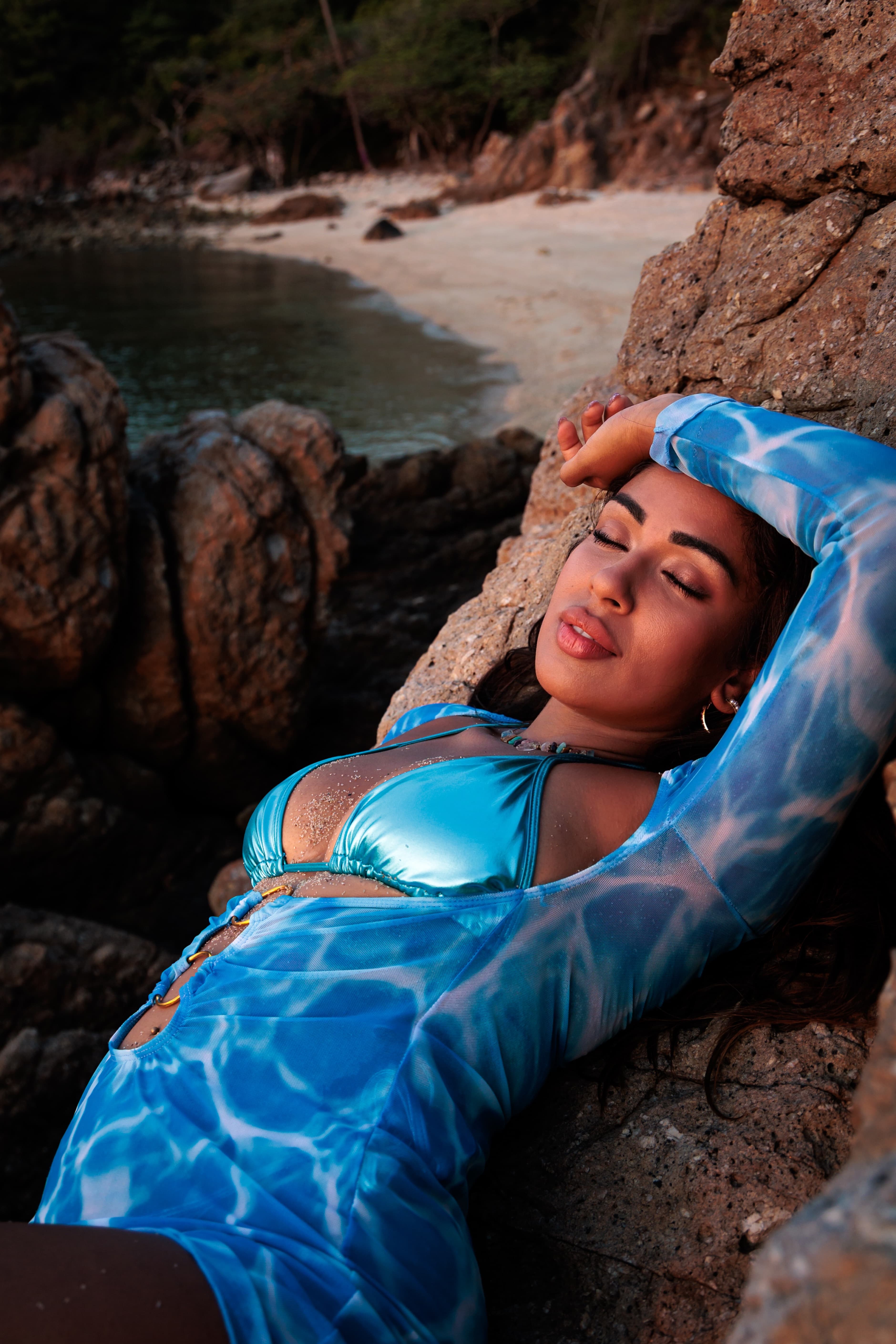 Frau im blauen Bikini liegt entspannt mit geschlossenen Augen auf Felsen an einem tropischen Strand.